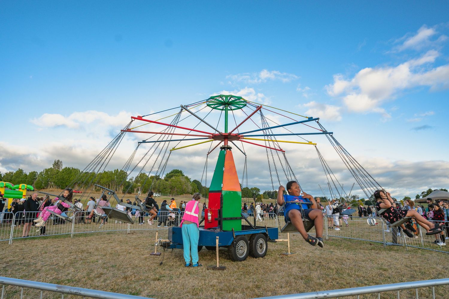 Colorful maypole carnival ride with people seated around it at an outdoor community festival.