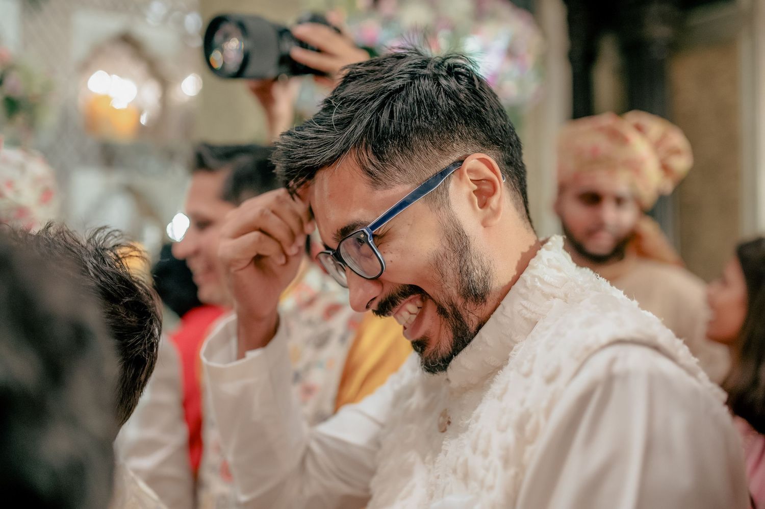 Laughing Indian groom in white sherwani adjusts glasses during joyful wedding celebration moment.