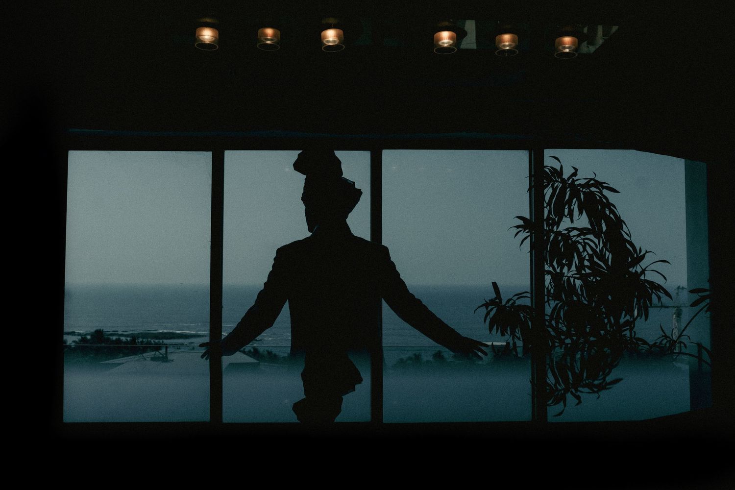 A dramatic silhouette of a groom in a turban standing before large ocean-view windows.