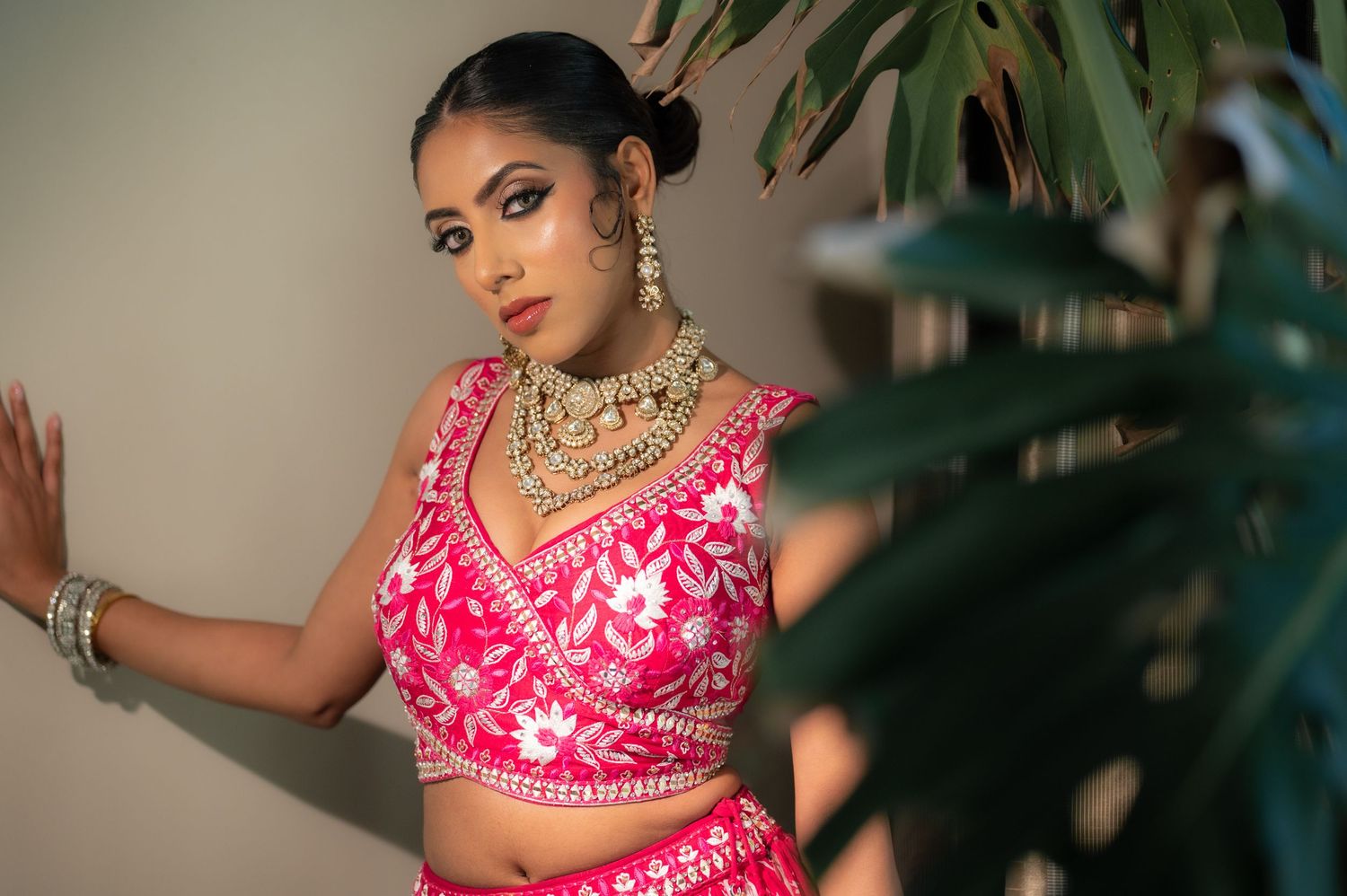 South Asian bride in vibrant pink lehenga with gold jewelry poses beside tropical plant.