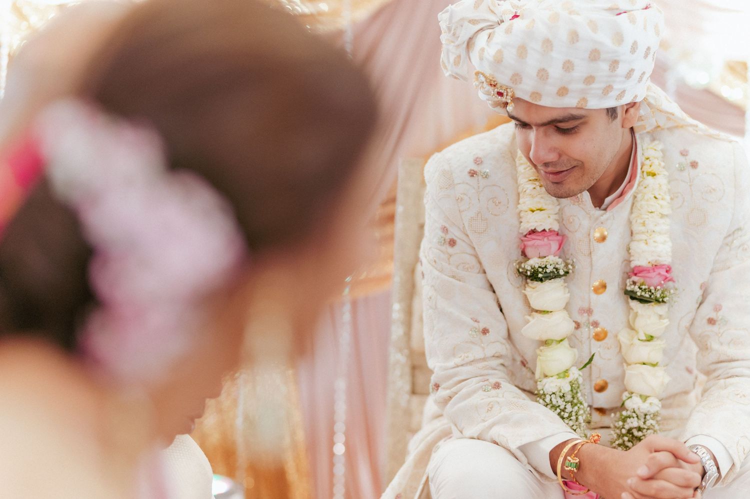Groom in cream sherwani and floral garland sits thoughtfully during Indian wedding ceremony rituals.