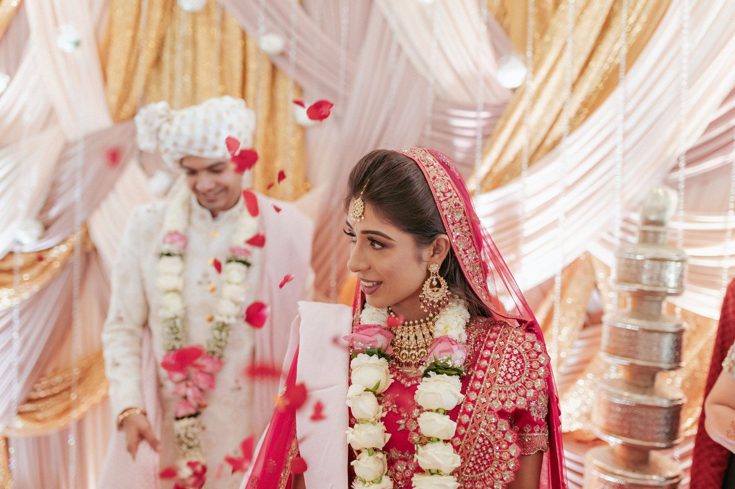 Elegant Indian bride in crimson lehenga with gold jewelry walks during traditional Hindu wedding ritual.