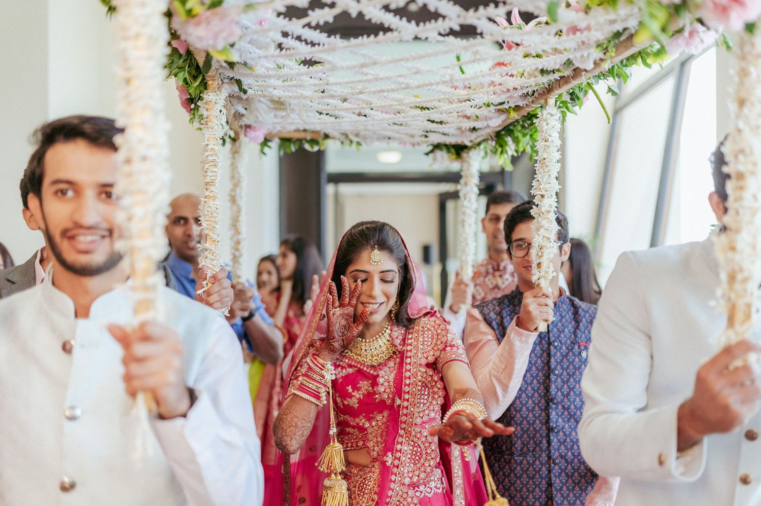 Indian bride in red lehenga makes grand entry under decorated floral wedding canopy.