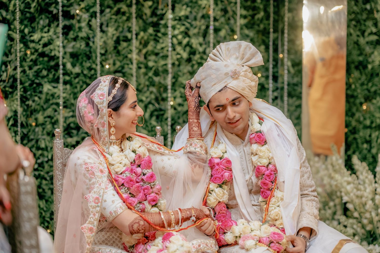 Indian bride and groom in white outfits with floral garlands share a joyful moment.