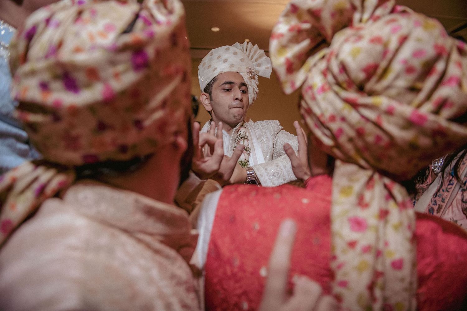 Indian groom in white sherwani and floral turban surrounded by groomsmen during wedding ceremony.