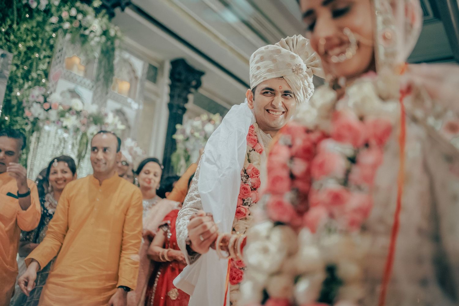 Groom in white sherwani and turban walks with bride during Indian wedding ceremony surrounded by guests.