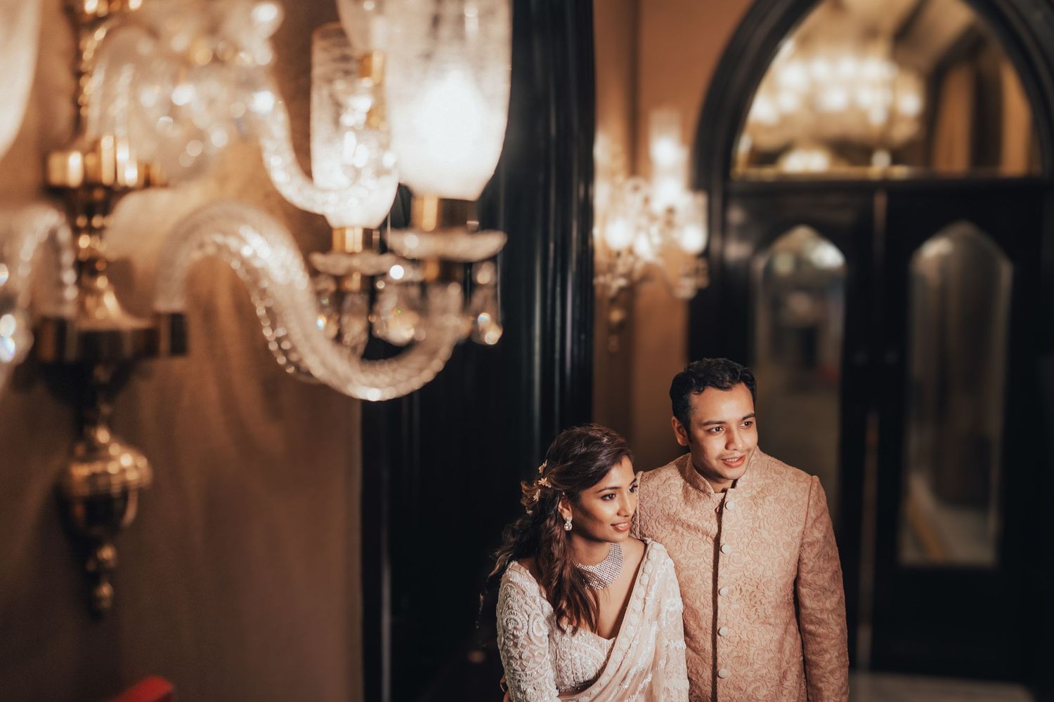 Elegant bride and groom in traditional Indian wedding attire pose near ornate crystal chandeliers indoors.