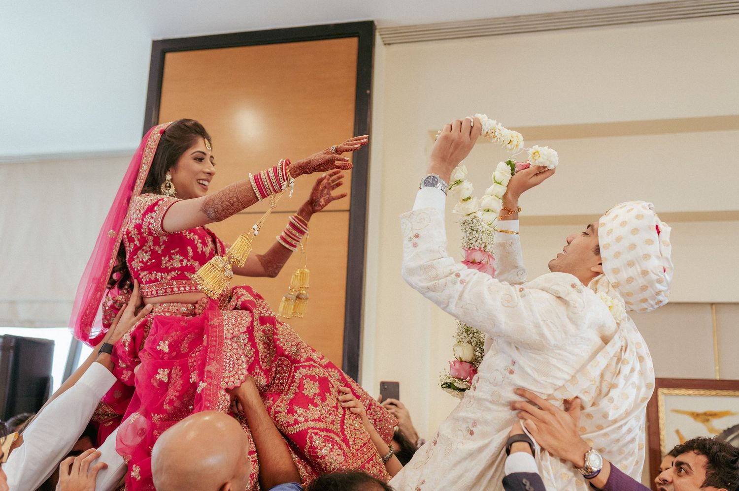 Indian bride in red lehenga reaches for garland as groom in white sherwani lifts it playfully.