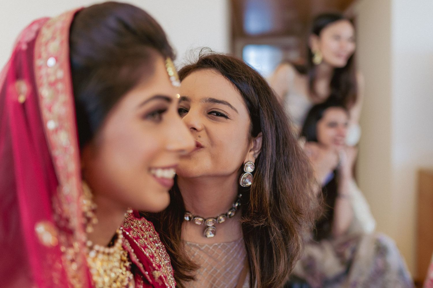 Female guest affectionately kisses Indian bride in red bridal lehenga on the cheek.