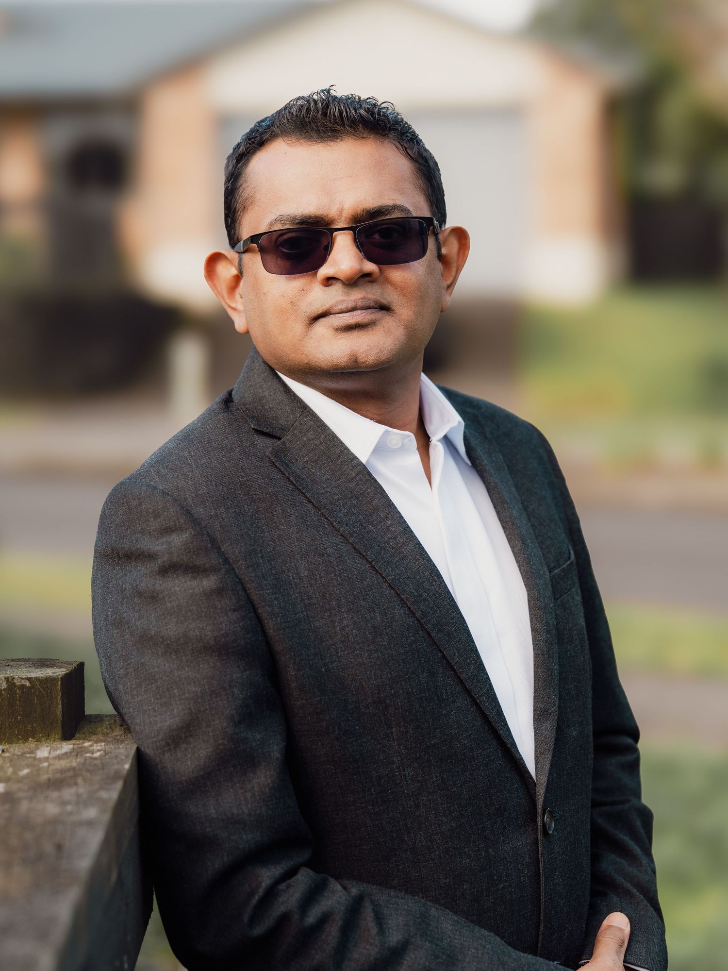 Man in dark blazer and sunglasses poses outdoors on a suburban street with a brick house behind him.