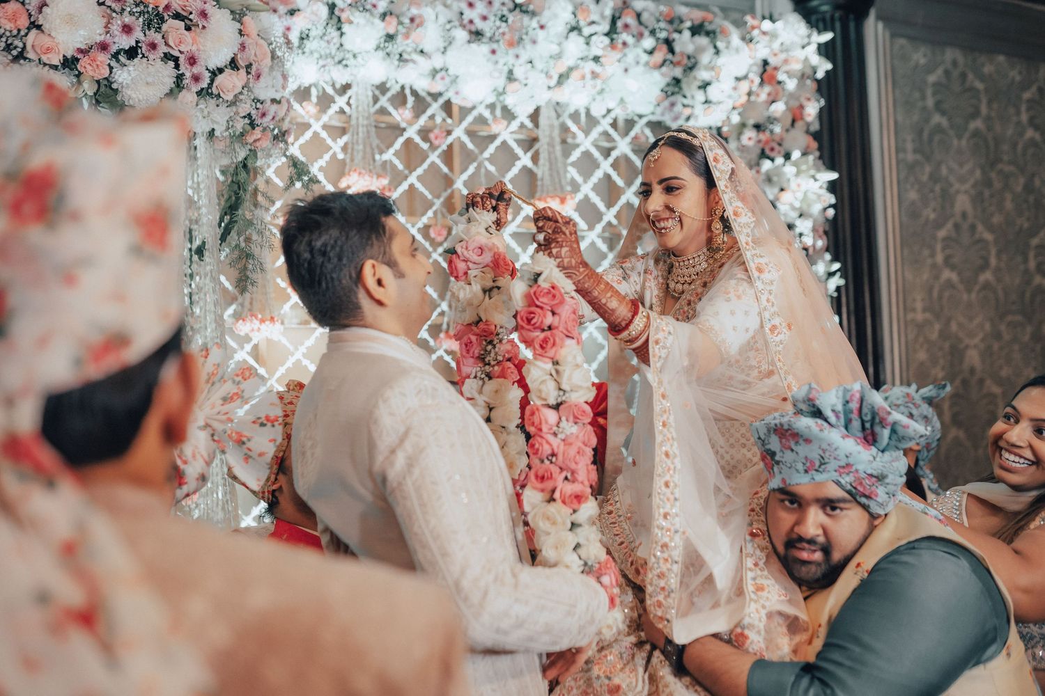 Indian bride and groom exchanging floral garlands at decorated wedding altar with pink flower backdrop.
