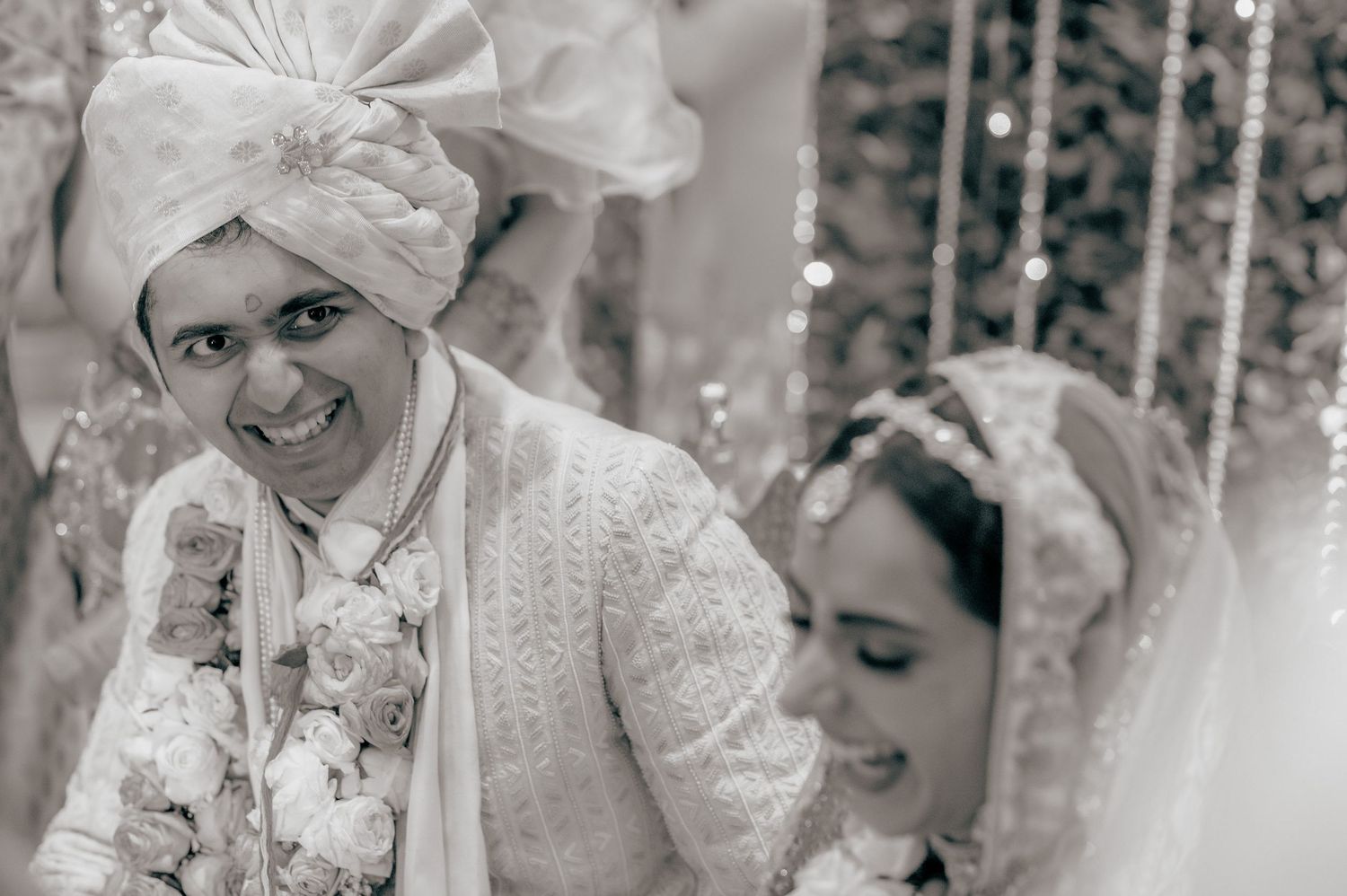 Smiling groom in white sherwani and turban with bride during black and white Indian wedding photo.