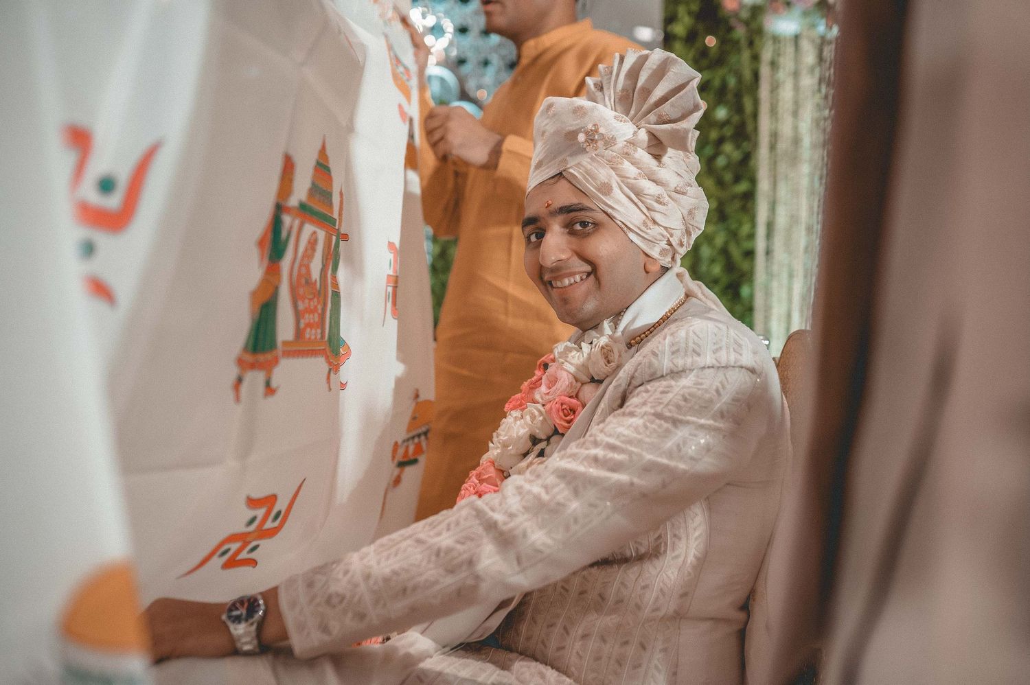 Smiling Indian groom in cream sherwani and turban sitting at traditional Hindu wedding ceremony.