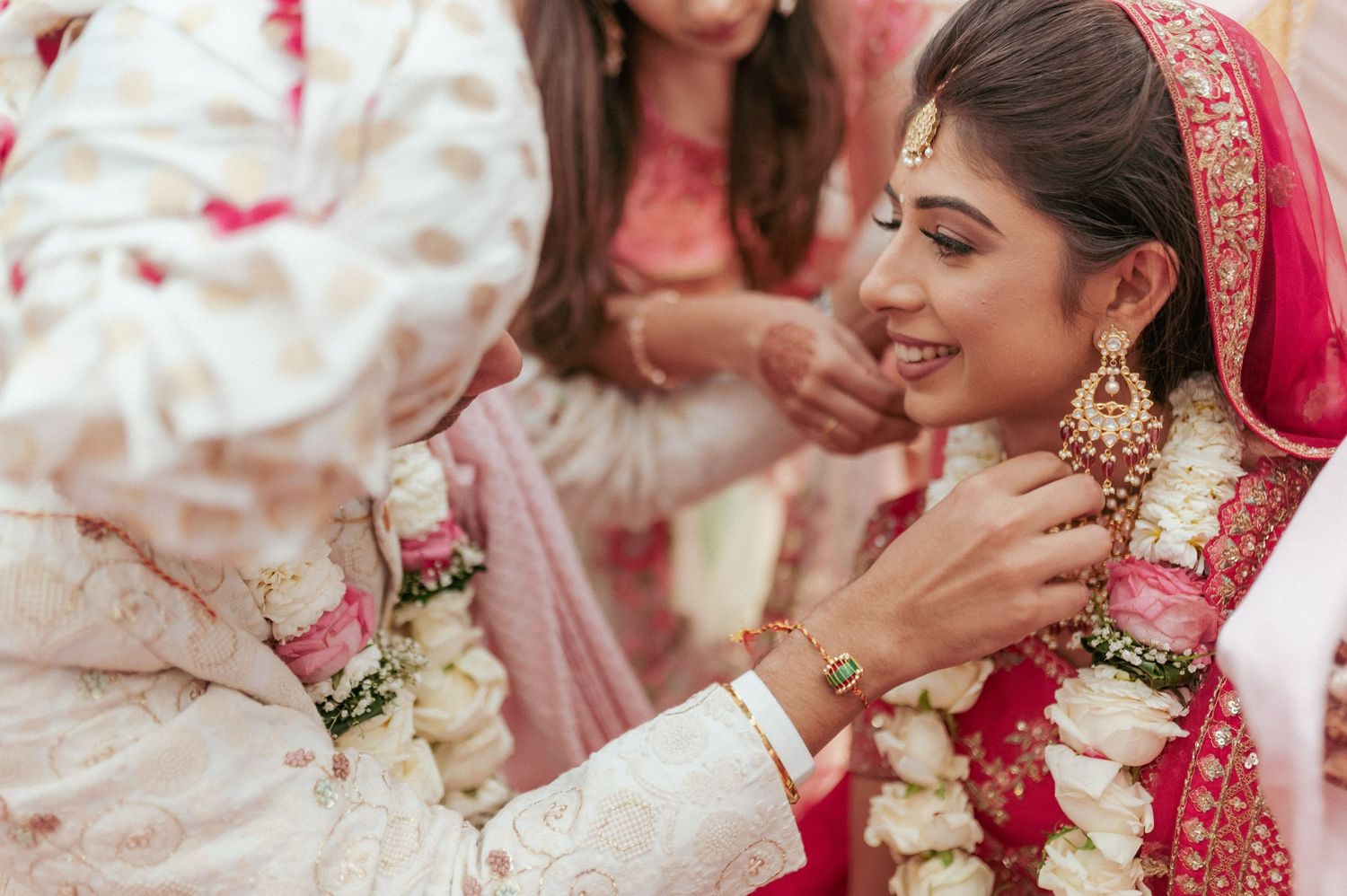 Indian bride in red lehenga and gold jewelry smiles as groom places garland during wedding ceremony.