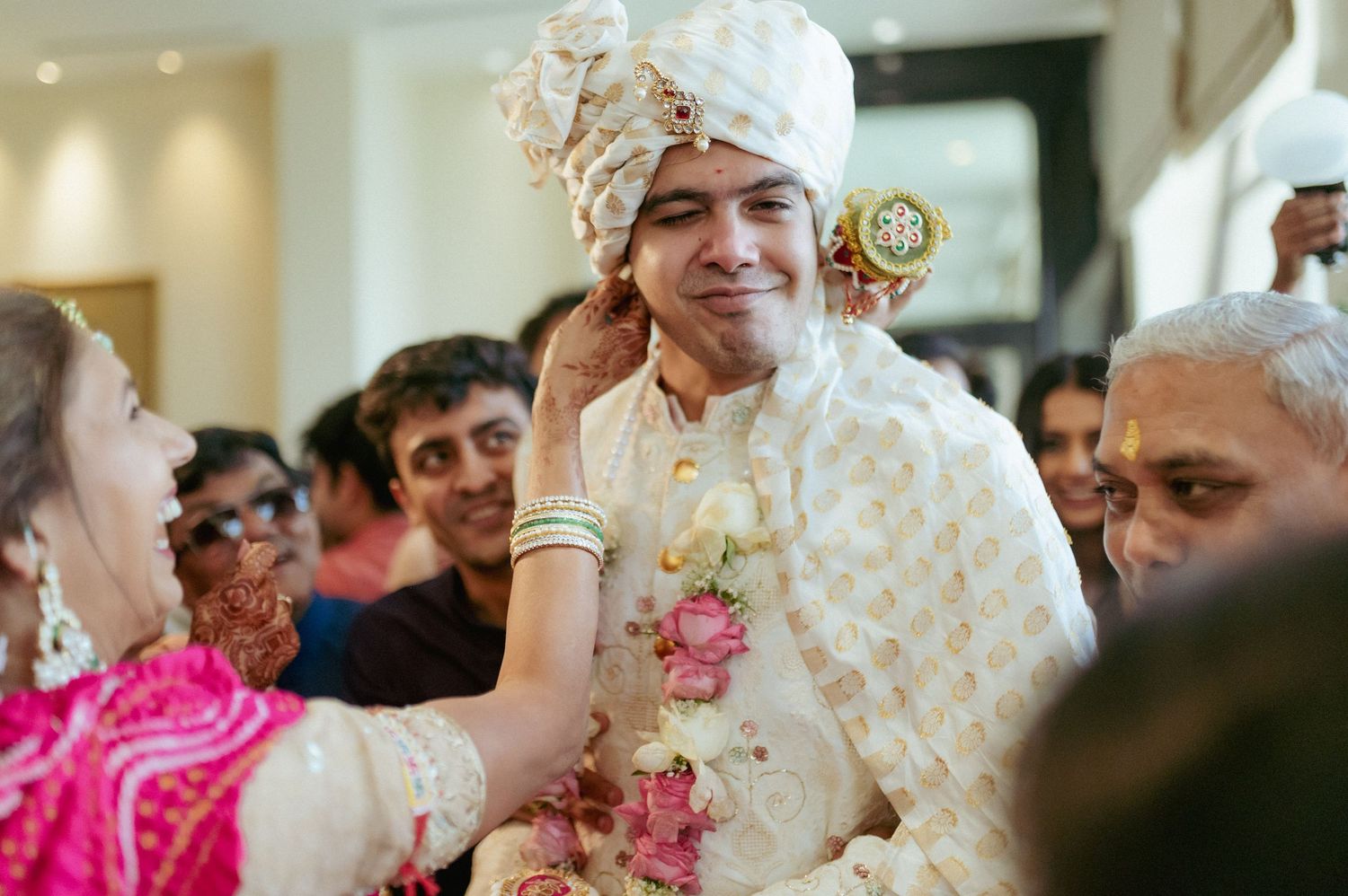 Groom in white sherwani and turban smiles as woman applies tilak during Indian wedding ceremony.
