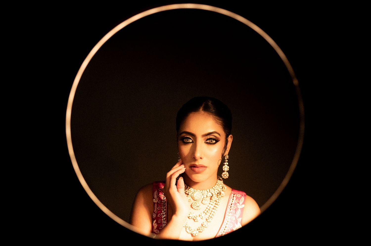 Elegant South Asian bride in pink saree with gold necklace framed within circular hoop in dark setting.