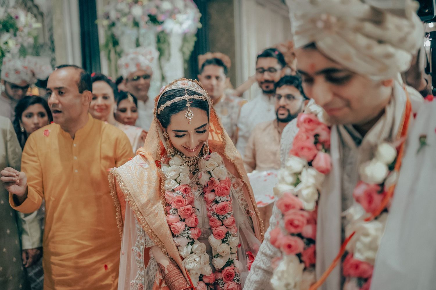 Indian bride in peach lehenga with floral garland walks through wedding aisle surrounded by family.