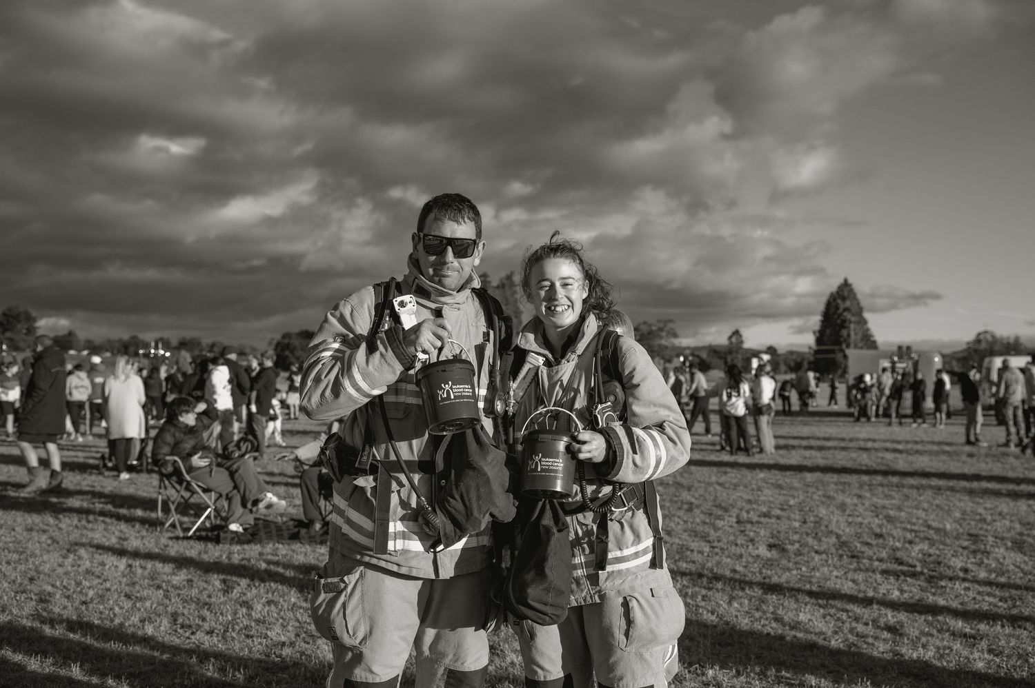 Black and white photo of two people in gear smiling at an outdoor festival with crowd behind them.