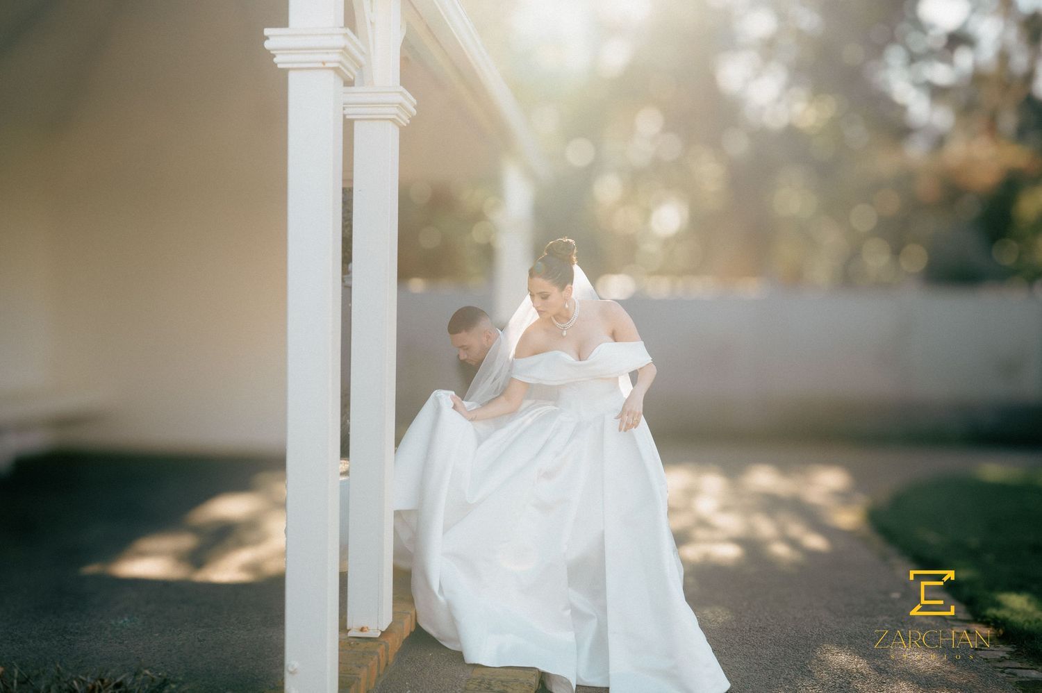 Groom carrying bride in white wedding dress through sunlit outdoor venue with white columns.