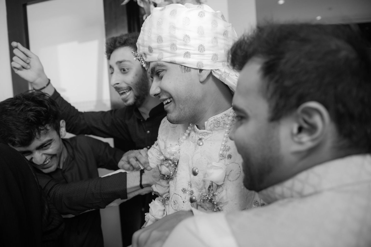 Groom in white sherwani laughs with friends in black and white Indian wedding photo.