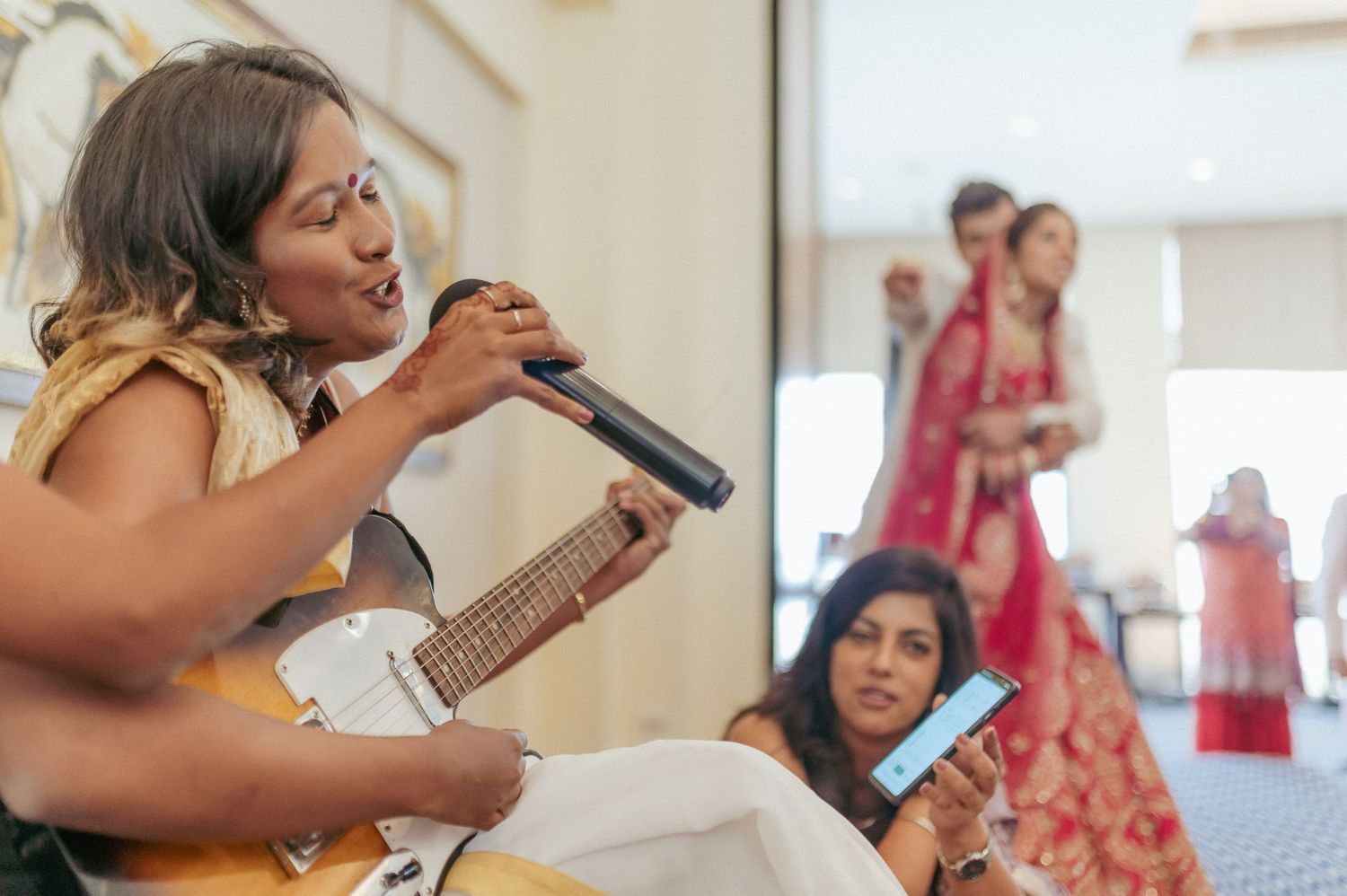 Live singer performing with microphone and guitar at Indian wedding with bride in background.
