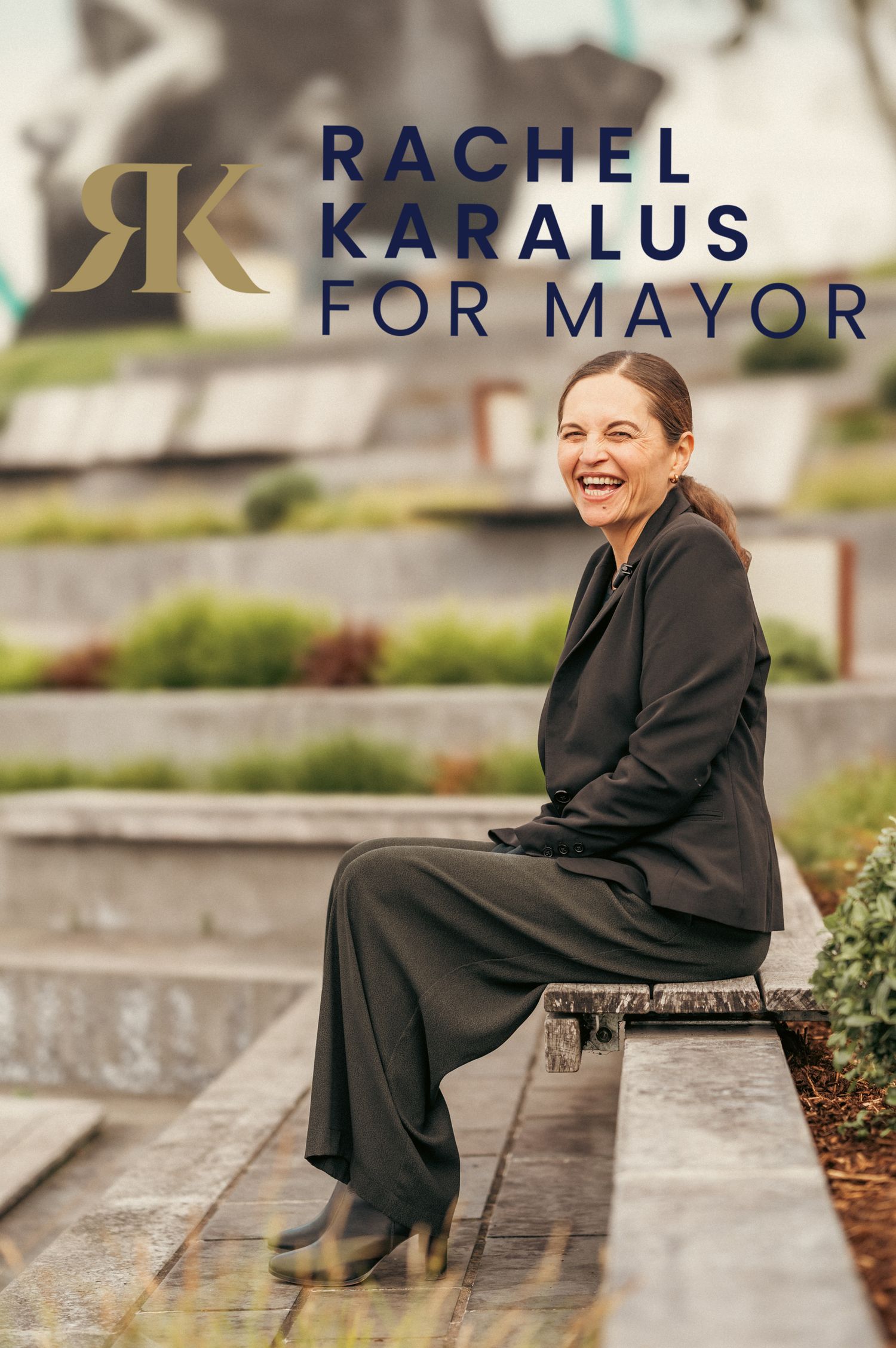 Rachel Karalus for Mayor campaign photo, smiling woman in black outfit seated outdoors near greenery.