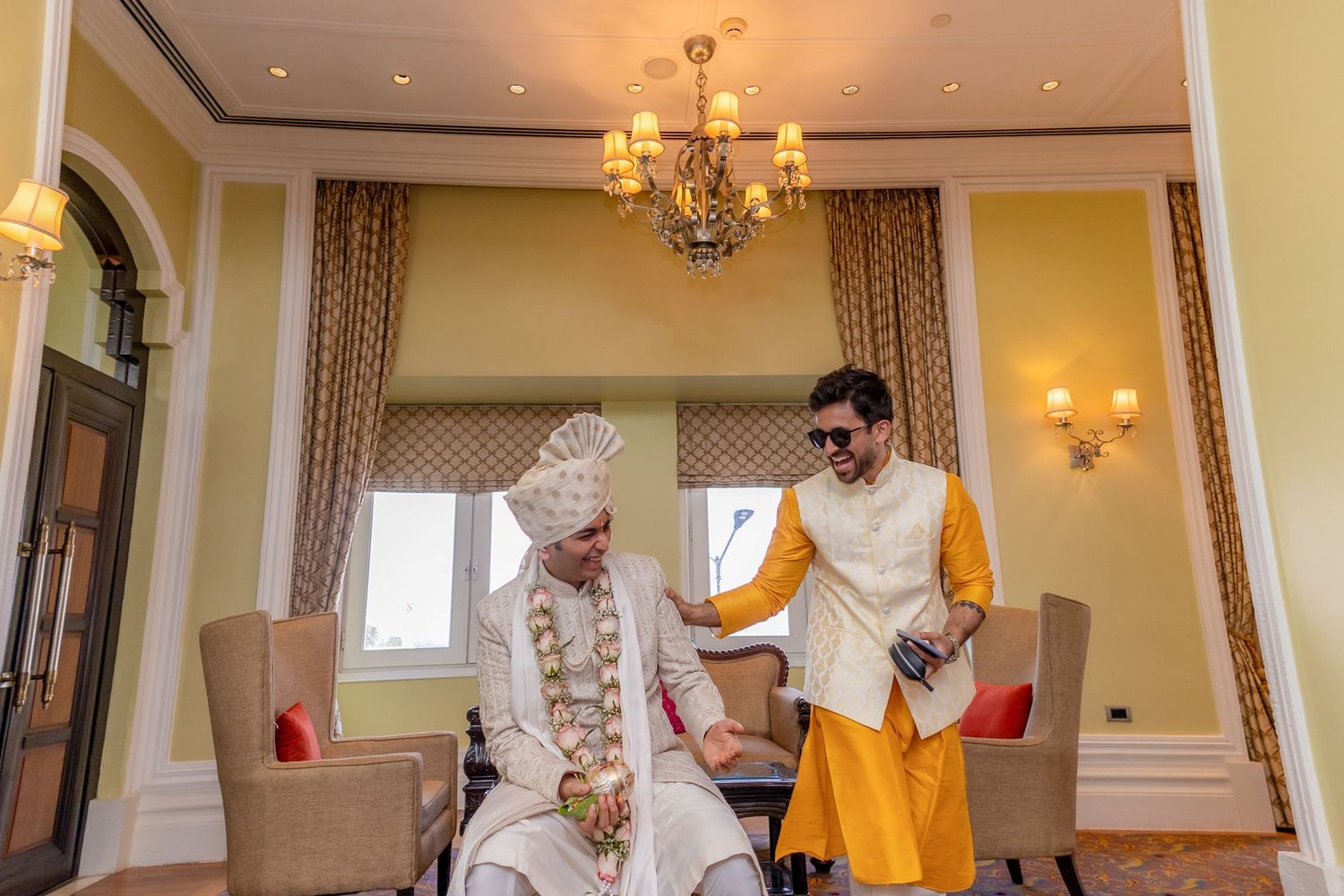 Two men in traditional Indian wedding attire share a moment in a luxurious hotel room.