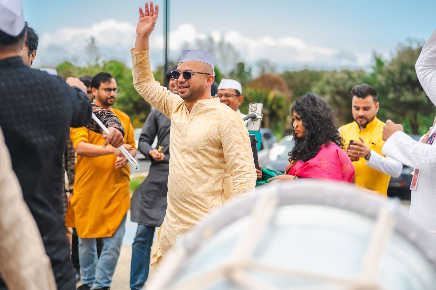 Man in gold kurta and sunglasses waves at an Indian celebration outdoors with guests in traditional attire.