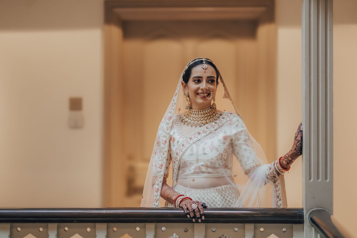 Indian bride in white embroidered lehenga and gold jewelry posing gracefully on a balcony railing.