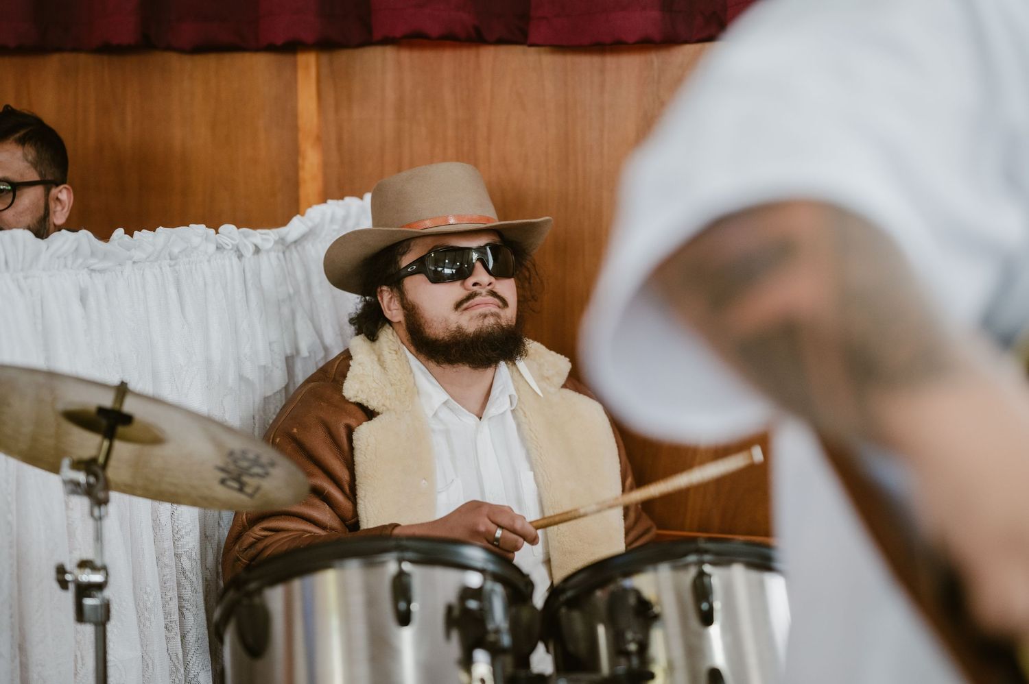 Musician in brown jacket and hat playing drums at an indoor religious or cultural music event.