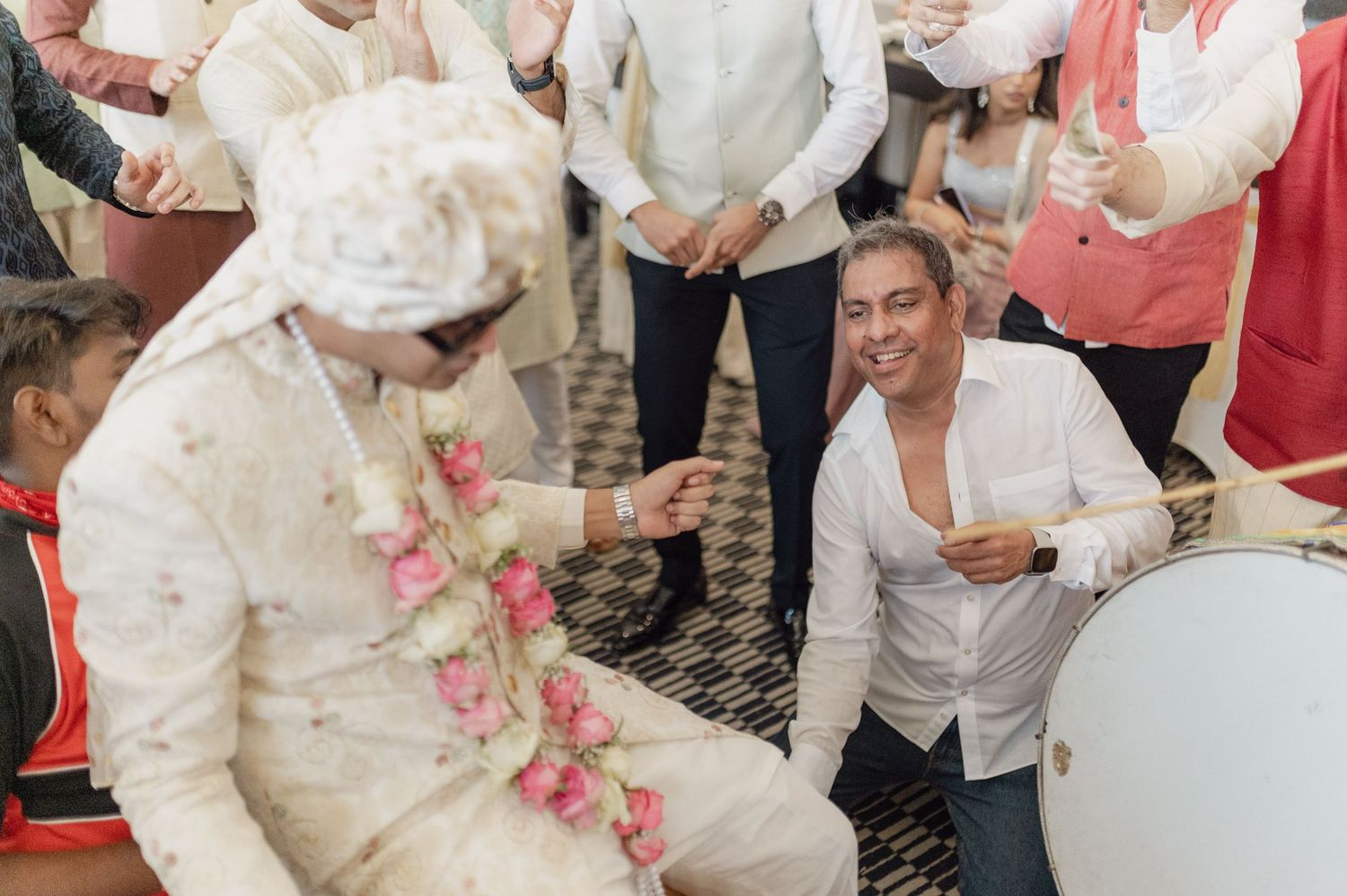 Groom in white sherwani with floral garland greeted by kneeling guest at Indian wedding ceremony.