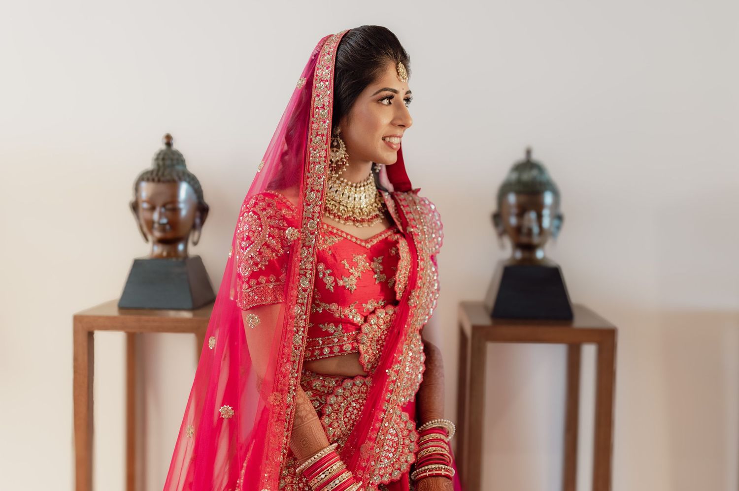 Elegant Indian bride in red embroidered lehenga with gold jewelry poses gracefully before wedding.