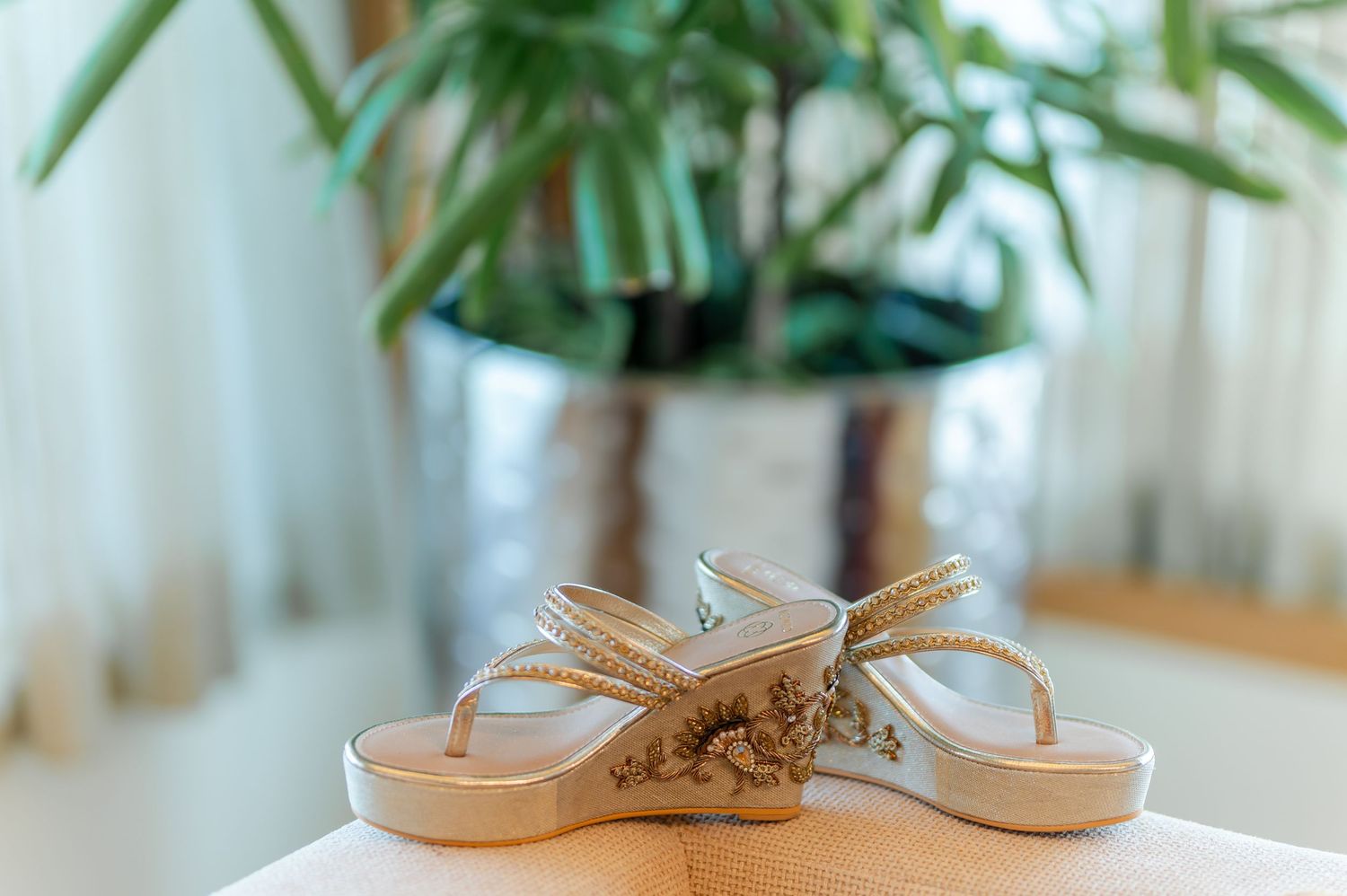 Embellished gold wedge sandals displayed on table with plant background.