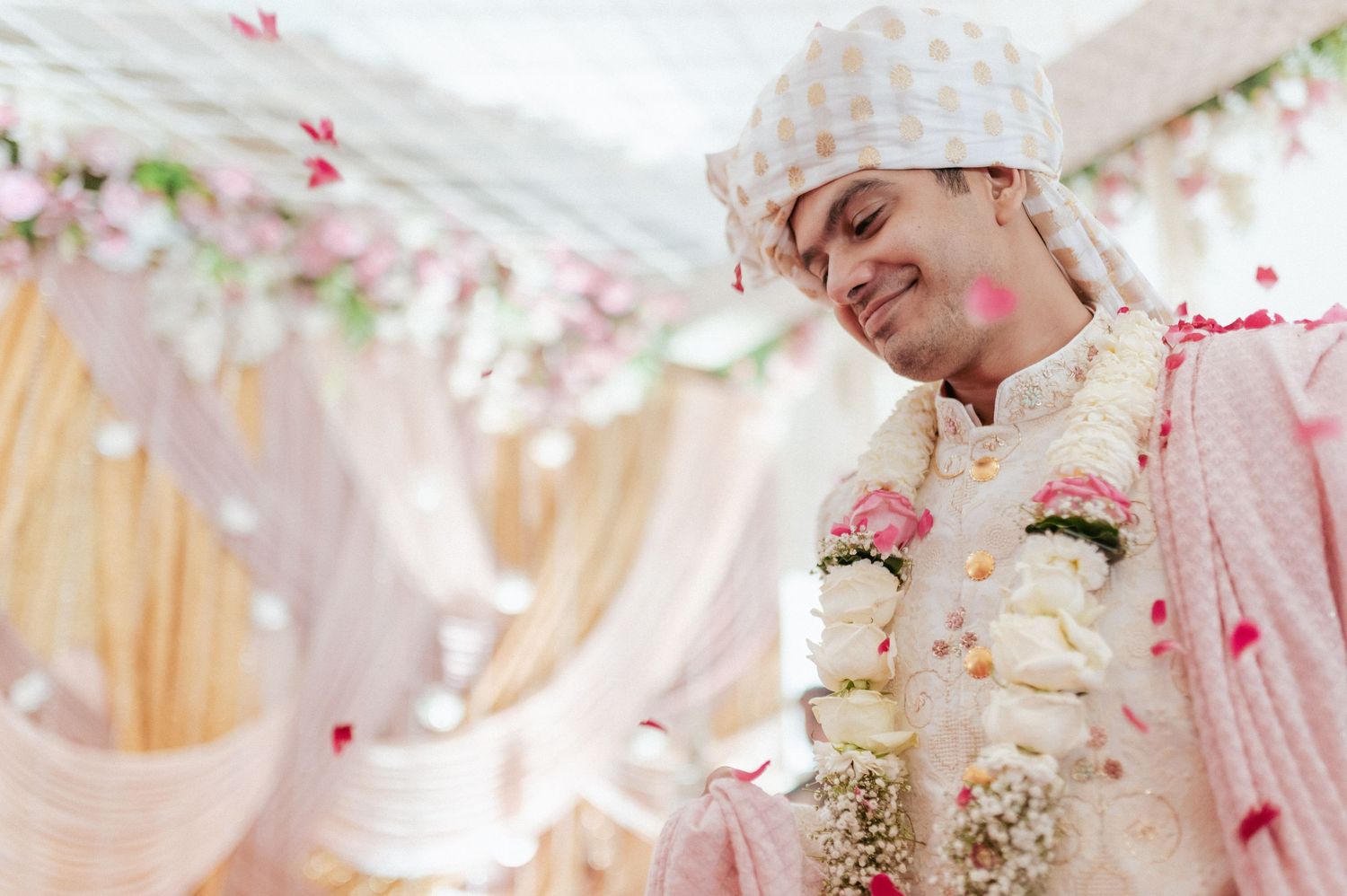 Handsome Indian groom in white sherwani and turban smiles as rose petals fall at wedding.