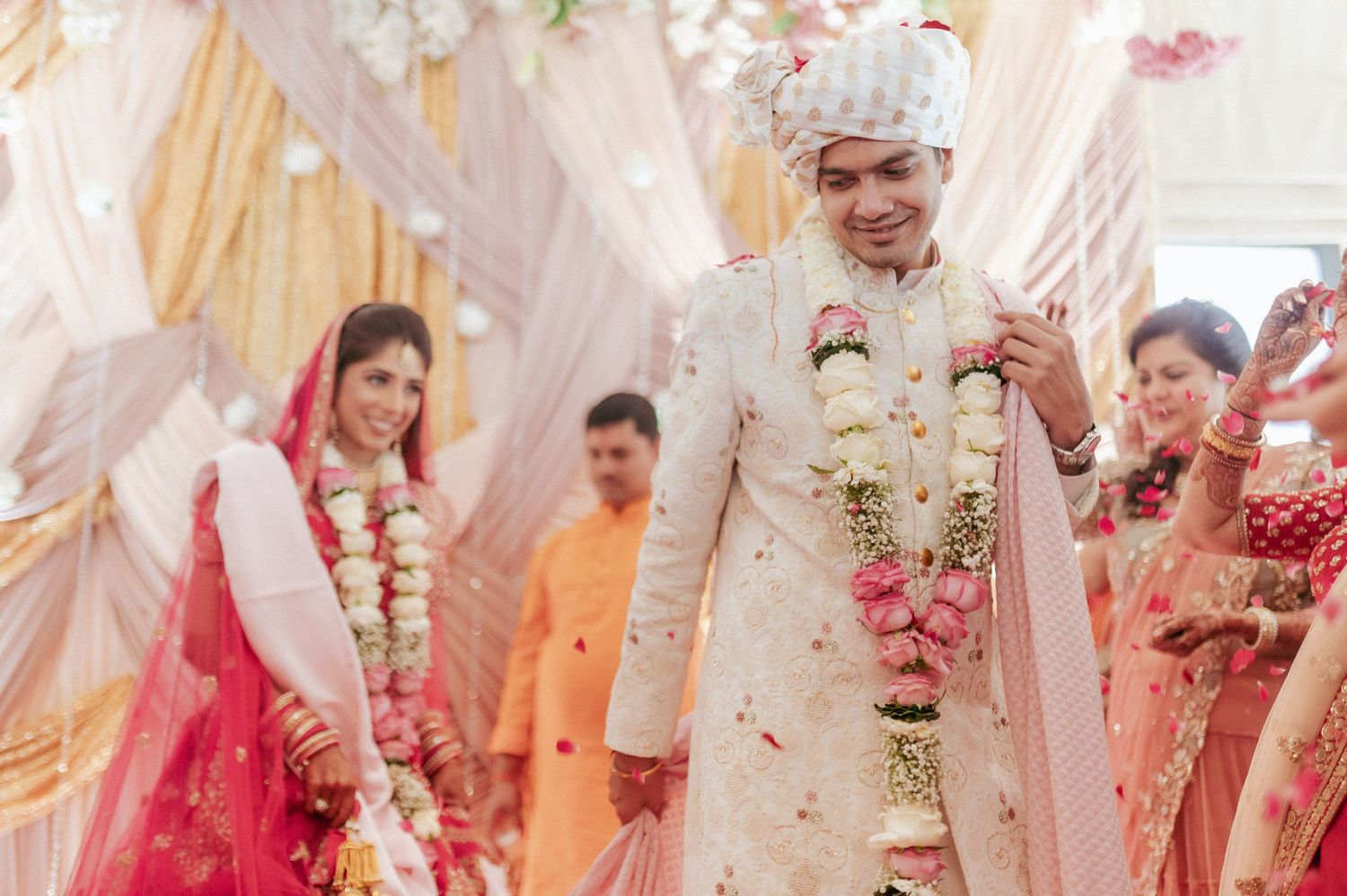 Smiling Indian groom in white sherwani holds floral garland while walking with bride at wedding.