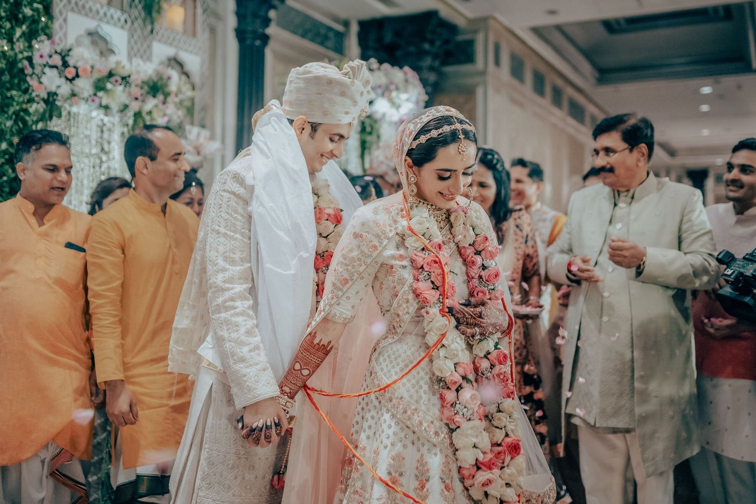 Indian bride and groom in white wedding attire walk hand in hand at ceremony.