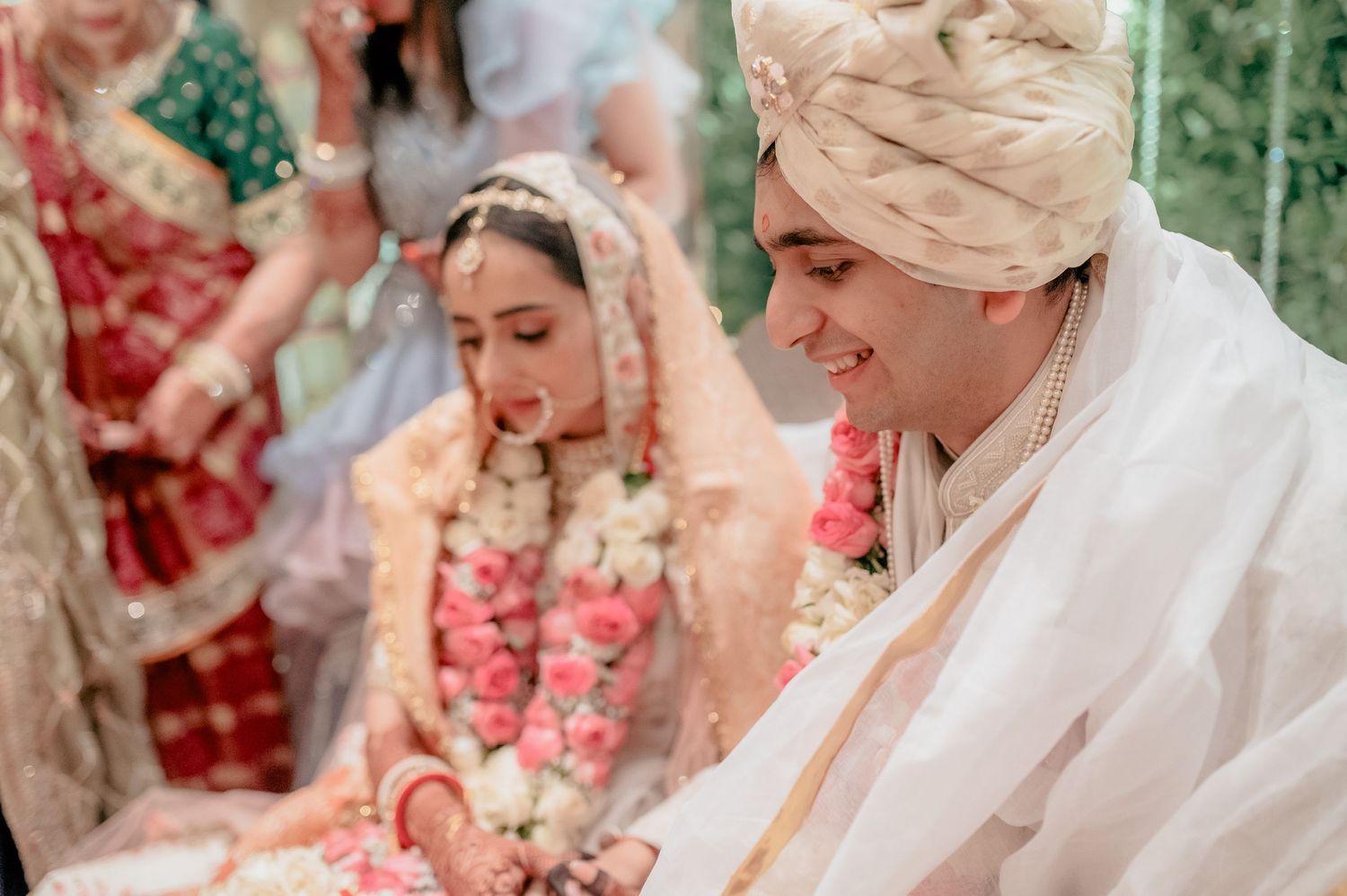 Indian groom in white sherwani sits beside bride adorned with pink rose garland during wedding rituals.
