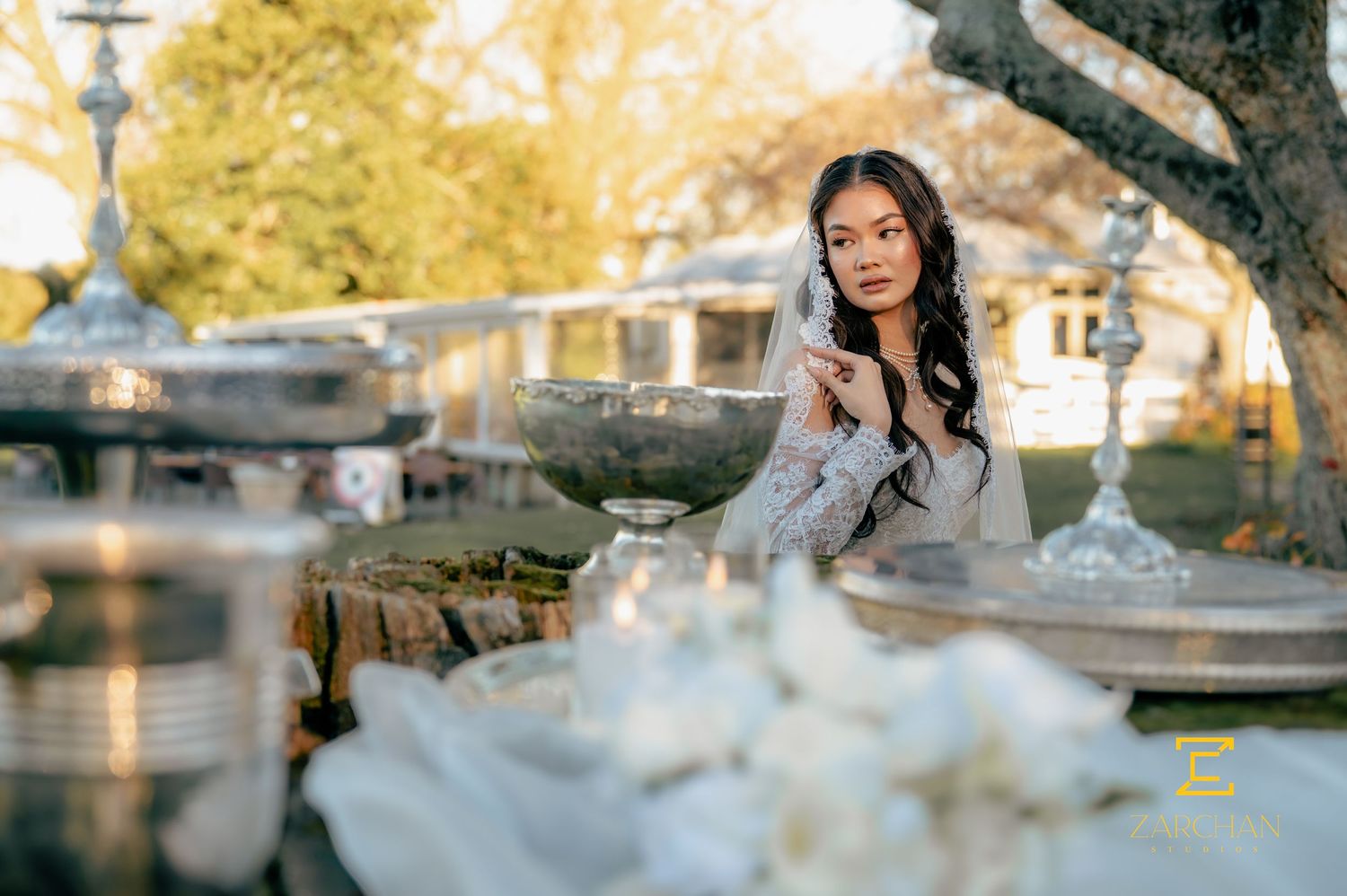Beautiful bride in lace wedding dress seated outdoors near elegant silver decorative fountains.