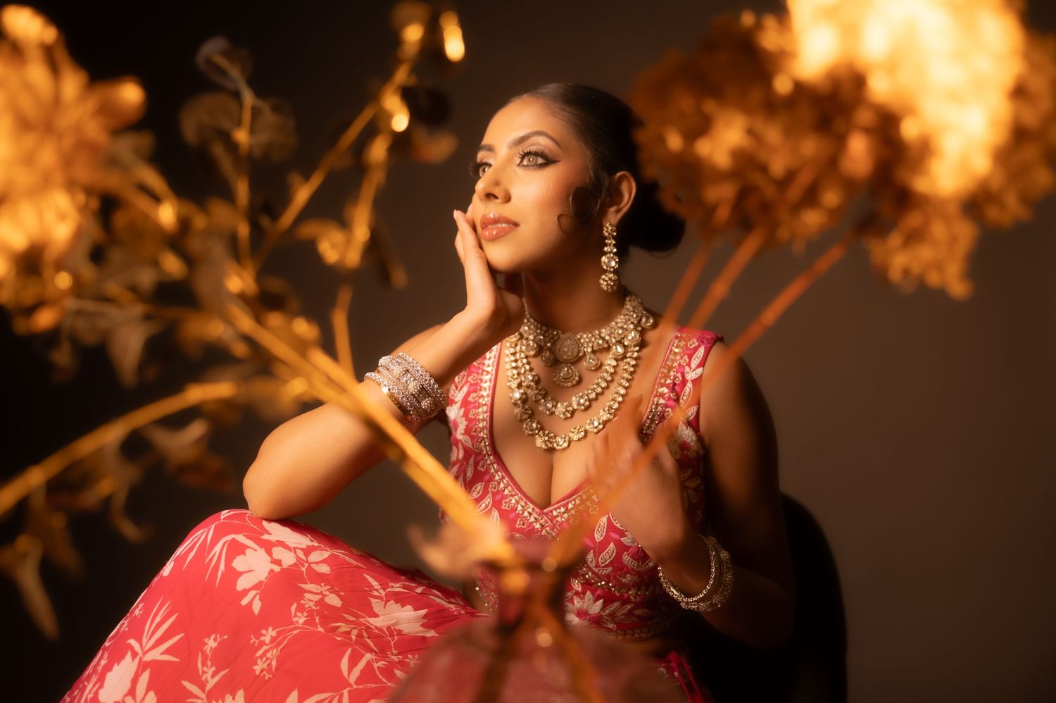 Woman in red floral lehenga with gold jewelry poses elegantly against warm golden backdrop.