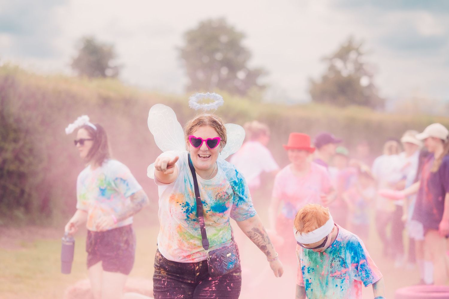 Woman in angel costume covered in colorful powder laughing during a fun color run event outdoors.