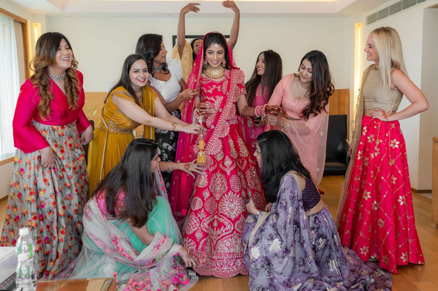 Bride in red lehenga surrounded by bridesmaids helping her prepare for Indian wedding ceremony.