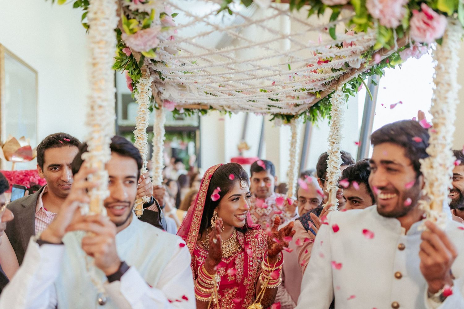 Bride in red lehenga walks under floral canopy at Indian wedding ceremony with smiling guests.