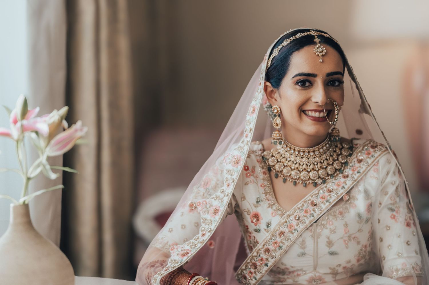 Smiling Indian bride wearing white lehenga with floral embroidery, gold jewelry, and bridal veil.