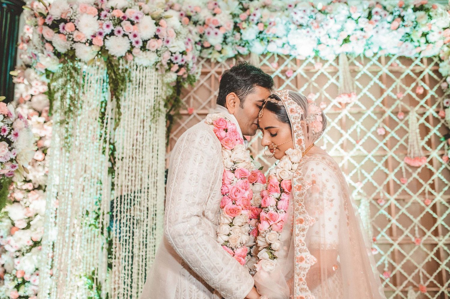 Indian newlyweds in ivory outfits sharing an intimate moment under a floral wedding mandap.