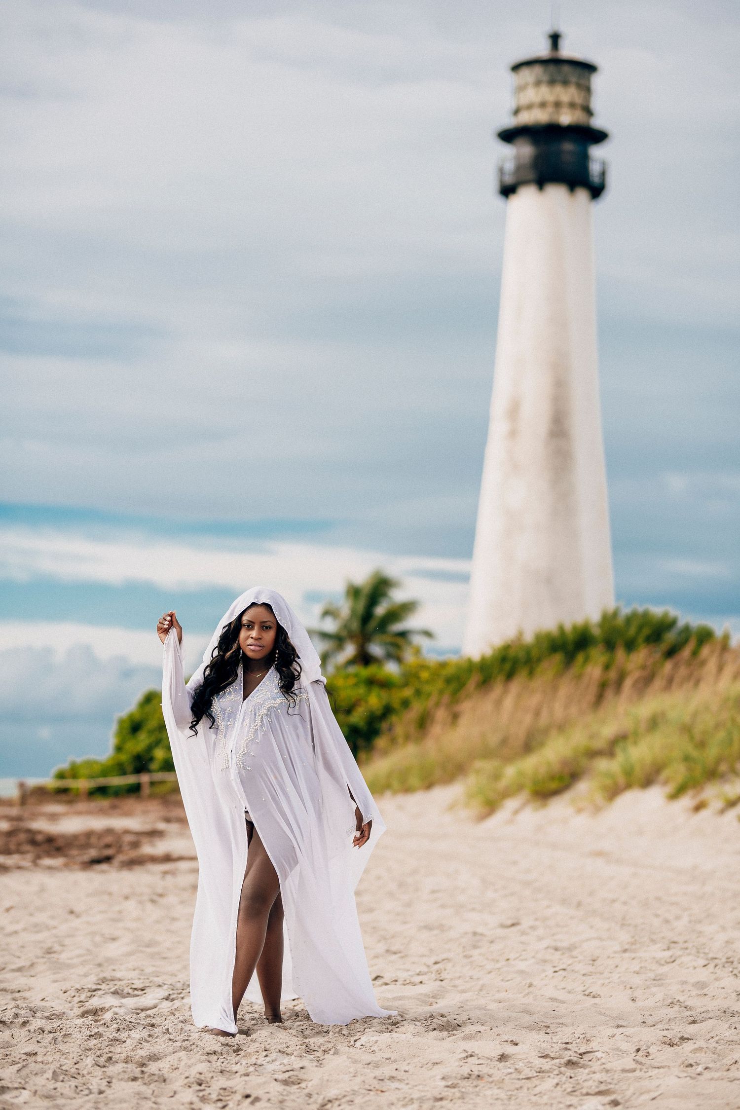 A figure in a flowing white dress dances gracefully on a sandy beach with a lighthouse in the background.