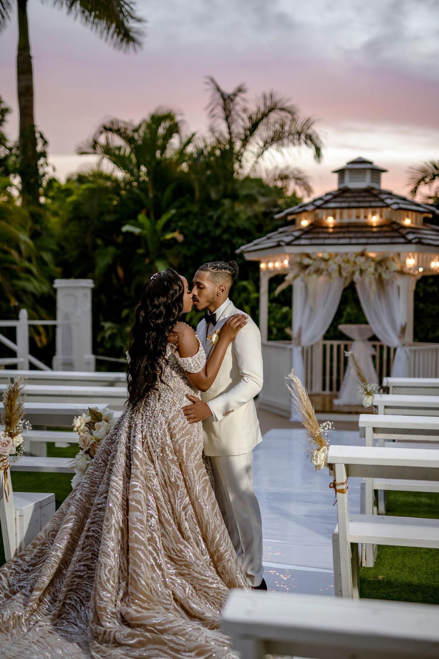 Romantic wedding ceremony at sunset with illuminated gazebo and palm trees in tropical garden setting.
