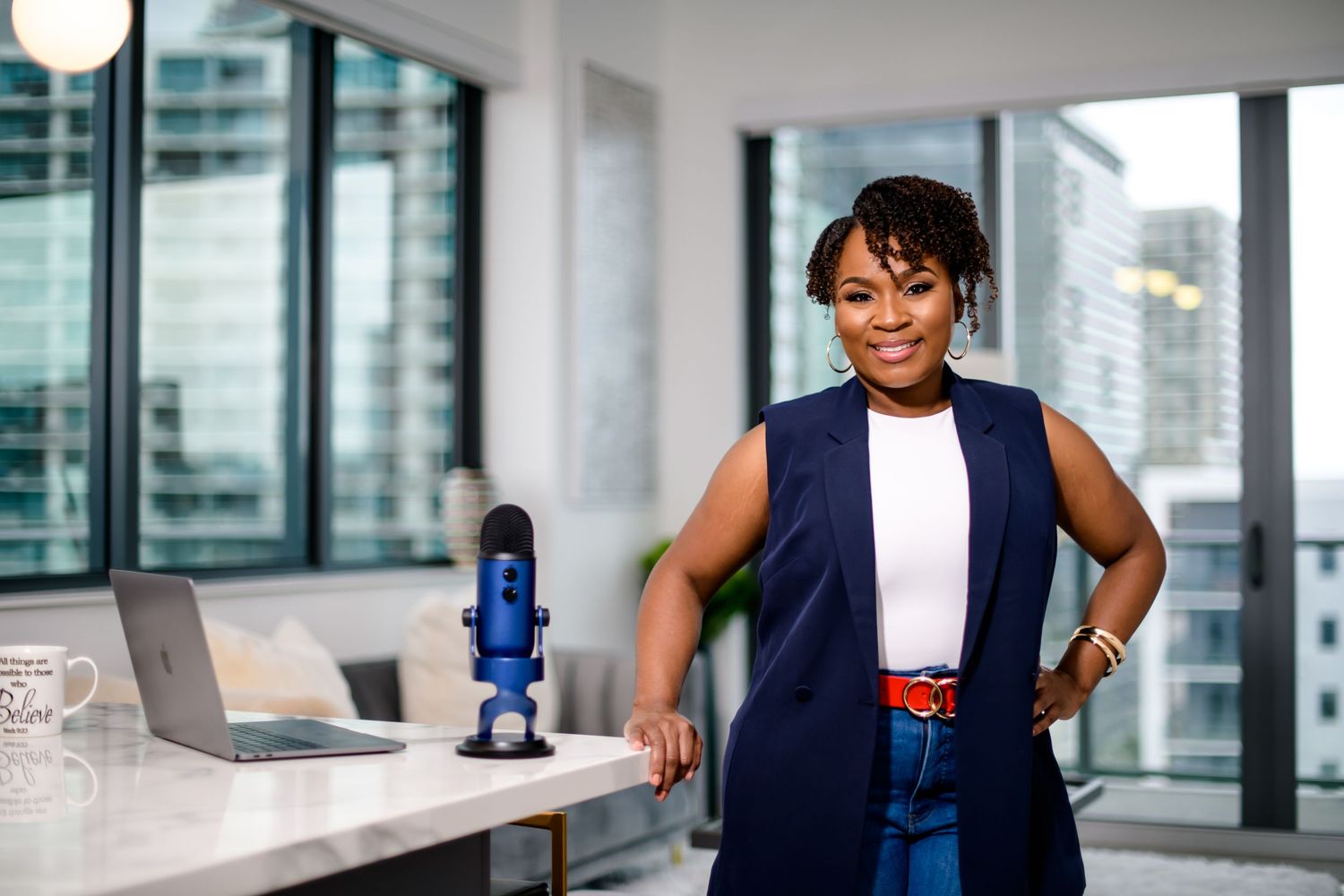 Professional woman in navy blazer poses confidently in modern office with Blue Yeti microphone and laptop on desk.