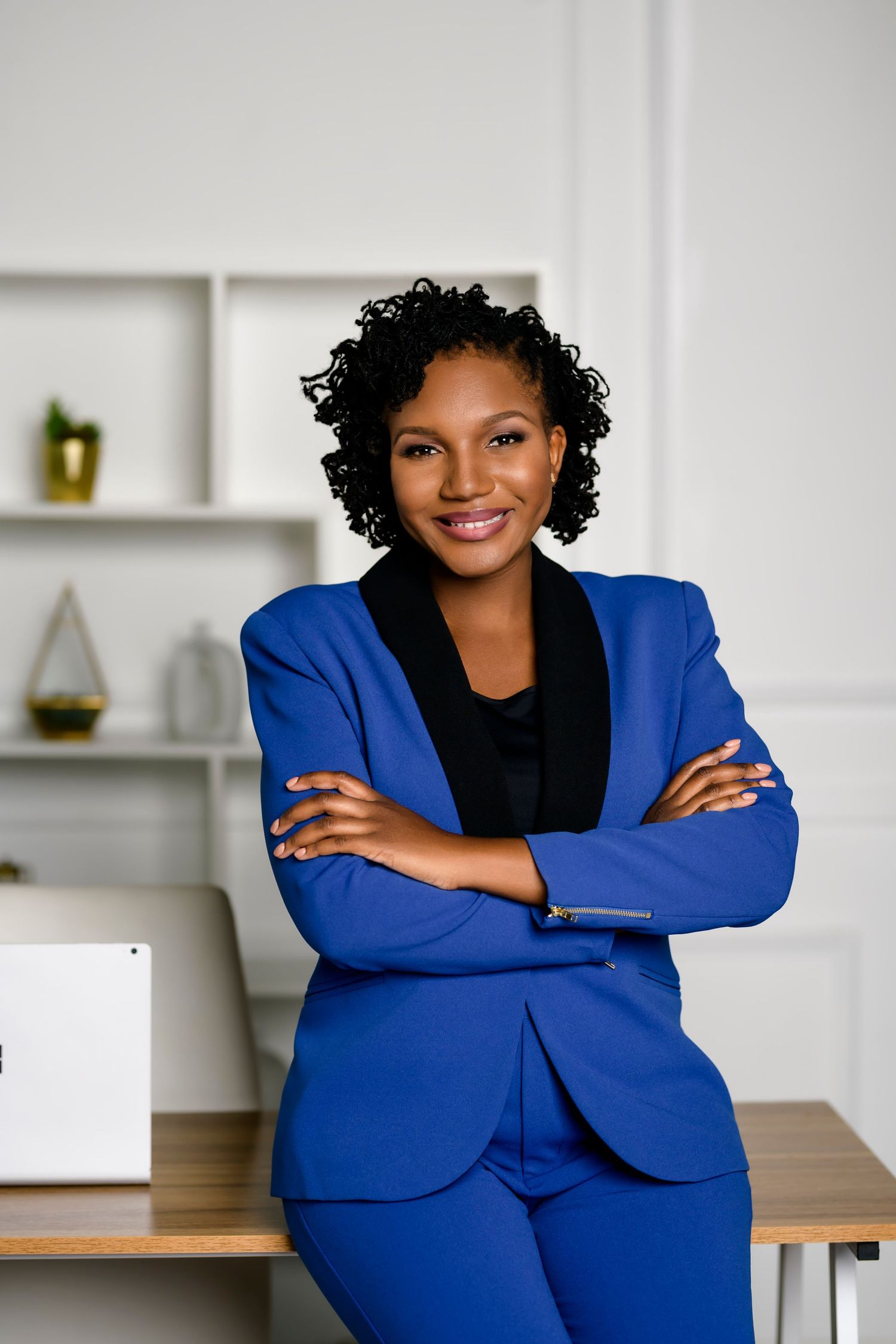 Professional Black woman in blue suit smiling with arms crossed, leaning on desk in modern office.
