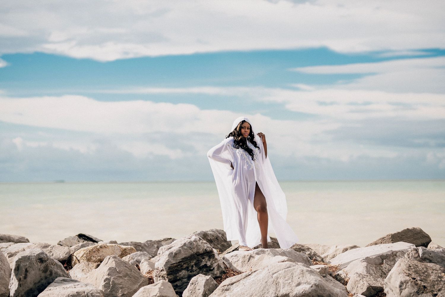A person in white dress walks along a rocky beach shoreline with blue sky and mountains in the background.
