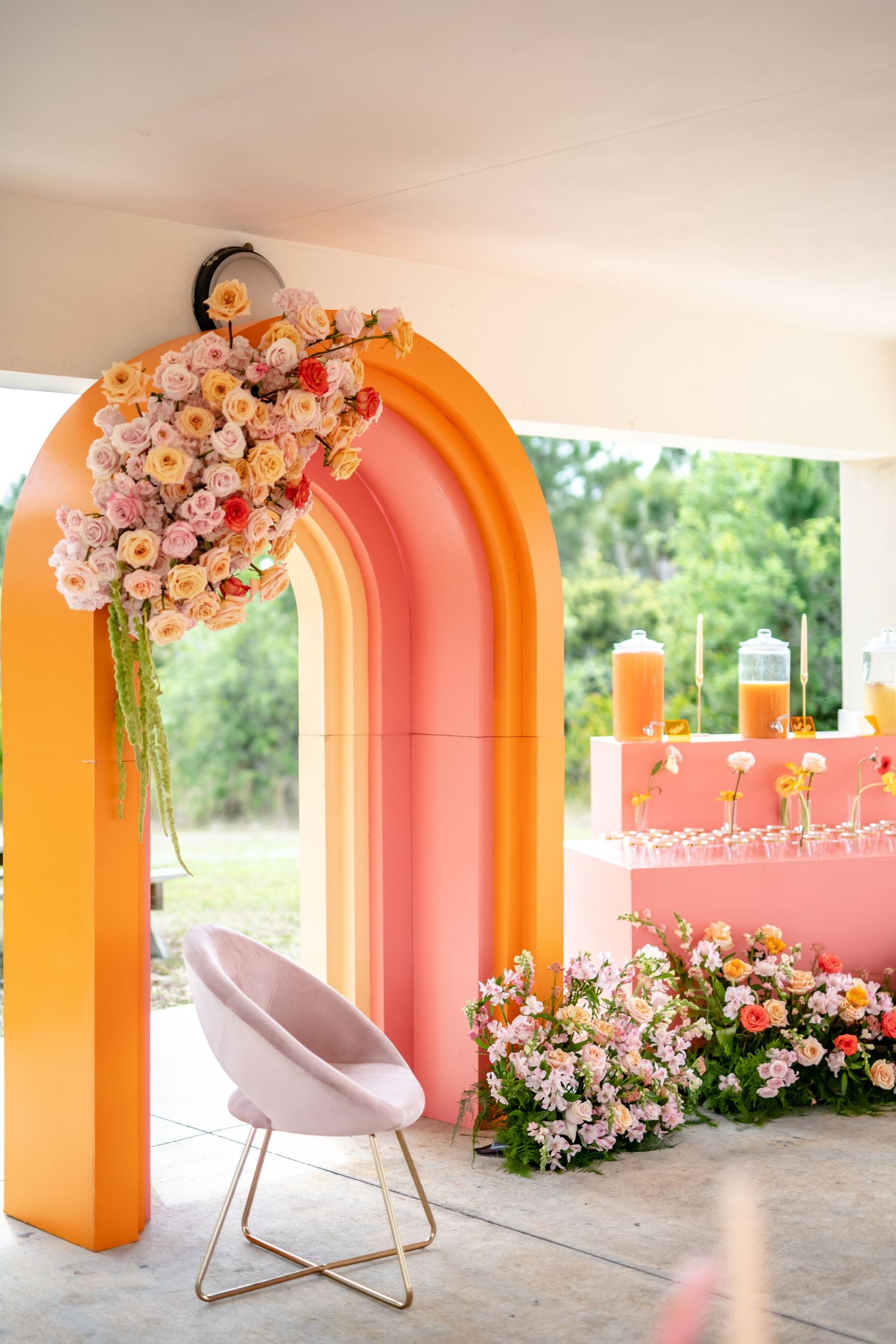 Modern event setup featuring a pink and orange gradient arch decorated with peach flowers and a plush velvet chair.